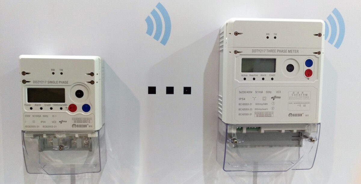 Two smart meters on a wall. One smart meter is a large white box with a red button and a blue button close to a small digital display.