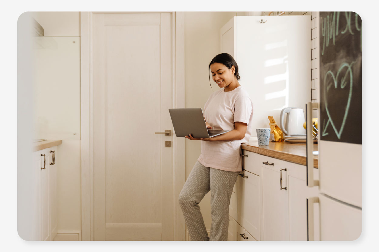 a woman stands in her kitchen on her laptop putting her bills into a bills package for one easy monthly payment