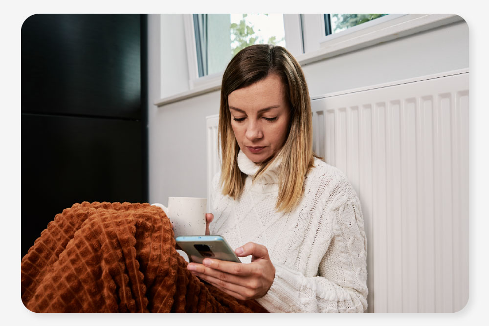 a woman sits cosy in a blanket looking skeptical at her phone. She's never heard of a One Utility Bill bills package before and wants to know why she should get one